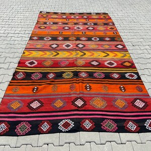 Pode incluir: Um tapete kilim turco vintage colorido com um padr&atilde;o geom&eacute;trico em tons de vermelho, laranja, amarelo, preto e rosa. O tapete tem uma forma retangular longa e &eacute; tecido com detalhes intrincados.