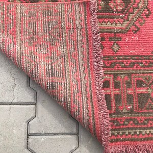 Puede incluir: Una alfombra de color rosa y marr&oacute;n con flecos en los bordes. La alfombra est&aacute; doblada por la mitad y se encuentra sobre una superficie de hormig&oacute;n gris.