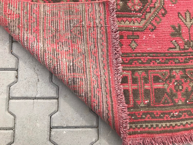 Puede incluir: Una alfombra de color rosa y marr&oacute;n con flecos en los bordes. La alfombra est&aacute; doblada por la mitad y se encuentra sobre una superficie de hormig&oacute;n gris.