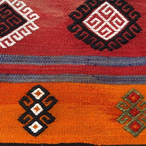 Pode incluir: Um close-up de um tapete tecido colorido com padr&otilde;es geom&eacute;tricos em tons de vermelho, laranja, azul, preto e branco.
