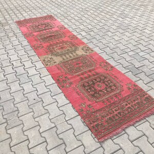 Puede incluir: Una alfombra larga y estrecha con un patr&oacute;n geom&eacute;trico rosa y marr&oacute;n. La alfombra tiene flecos en ambos extremos.
