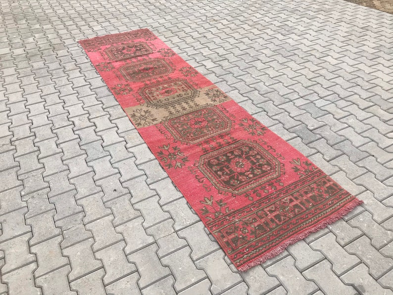 Puede incluir: Una alfombra larga y estrecha con un patr&oacute;n geom&eacute;trico rosa y marr&oacute;n. La alfombra tiene flecos en ambos extremos.