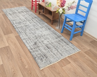 Puede incluir: Una alfombra de pasillo gris y blanca desgastada con un aspecto descolorido y vintage. La alfombra es rectangular, con un borde con flecos, y está colocada sobre un suelo de madera de color claro. El diseño de la alfombra presenta un patrón moteado, lo que se suma a su estética envejecida.