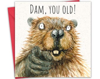 May include: A square greeting card featuring a watercolor illustration of a brown beaver with wide eyes, pointing forward with one paw. The text DAM, YOU OLD! is printed in black at the top. A red envelope is visible behind the card.