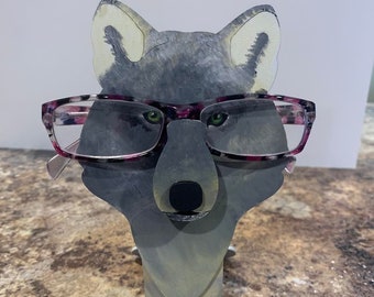 May include: A gray wolf-shaped wooden glasses holder with pink and black glasses perched on its nose. The wolf is standing on a brown speckled surface.