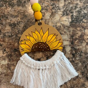 May include: A wooden sunflower cutout with a white macrame fringe hanging from the bottom. The sunflower is painted yellow with a brown center. There are three wooden beads in yellow and white hanging from the top.