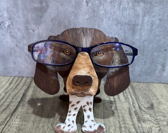 May include: A brown and white dog figurine wearing blue glasses. The figurine has a brown head, brown ears, and white paws. The figurine is standing on a wooden surface.