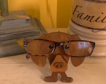 Może przedstawiać: Drewniany stojak na okulary w kształcie psa rasy dachshund, pomalowany na brązowo z czarnymi oczami. Stojak na okulary ma na sobie okulary w brązowych oprawkach i przezroczystymi szkłami.
