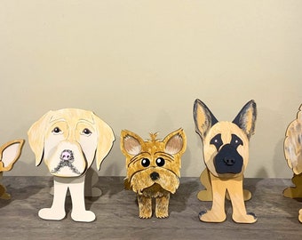 May include: A collection of seven wooden dog figurines, each painted with unique details. The figurines are standing on a wooden surface. The dogs are a Great Dane wearing glasses, a Chihuahua, a Labrador Retriever, a Yorkshire Terrier, a German Shepherd, a Poodle, and a unicorn.