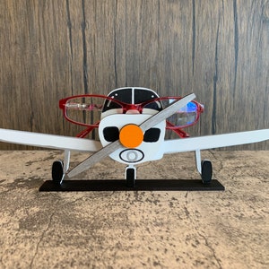 Puede incluir: Un soporte para gafas de madera blanca con forma de avión con un par de gafas rojas apoyadas en las alas. El avión tiene una base negra y una hélice naranja.