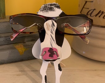May include: A black and white cow-shaped glasses holder with a pink nose and brown glasses. The holder is made of paper and has a painted design.