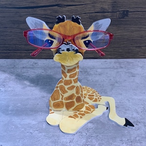 May include: A wooden giraffe figurine with a brown and yellow spotted pattern, wearing red glasses. The giraffe is sitting with its legs crossed and its head tilted to the side. The glasses have a red frame and clear lenses. The figurine is a decorative item for a home or office.