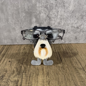 Könnte beinhalten: Ein Brillenhalter in Form eines Hundes aus Holz. Der Hund hat ein schwarz-weißes Gesicht, graue Beine und einen cremefarbenen Bart. Klare Brillen sind auf dem Gesicht des Hundes platziert. Der Halter steht auf einer Holzoberfläche.