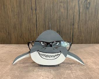 May include: A gray wooden shark with a toothy grin and black glasses. The shark is a decorative stand for eyeglasses.