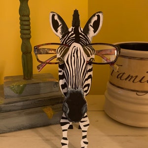 Puede incluir: Una figurita de cebra en blanco y negro con gafas rosas y transparentes. La cebra está de pie sobre una superficie blanca.