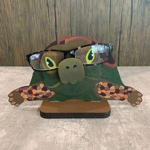 May include: A wooden turtle-shaped eyeglasses holder with a brown base. The turtle is wearing black framed glasses with green lenses and has a green, brown, and orange shell.