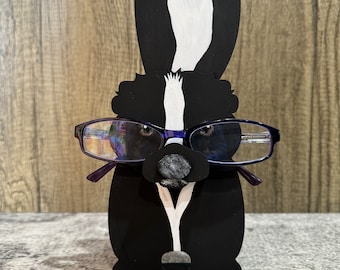 May include: A black and white wooden skunk figurine with a gray felt nose, wearing blue glasses, and standing on a black base. The skunk is holding the glasses in its mouth.