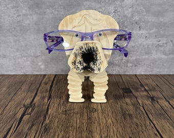 May include: A wooden dog sculpture with a light brown finish, wearing purple-framed glasses. The dog has a detailed, textured snout and is standing on a wooden surface. The background is a gray wall.