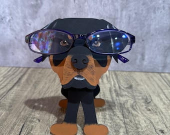 Może przedstawiać: Figurkę psa rasy Rottweiler w kolorze czarnym i brązowym, w niebieskich okularach. Figurka stoi na drewnianej powierzchni.