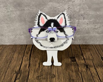 May include: A white, black, and pink husky dog-shaped eyeglasses holder with purple glasses. The holder has a detailed painted design of a husky dog's face and ears. The holder stands upright on two white feet.