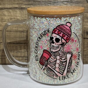 May include: A glass mug with a bamboo lid, filled with iridescent glitter. The mug features a skeleton graphic wearing a pink beanie and holding a red cup. Text on the mug reads "It's cold outside like my heart."