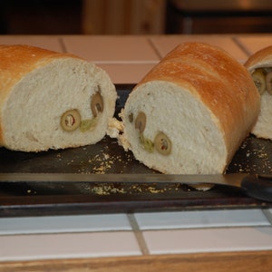 Könnte beinhalten: Drei Scheiben knuspriges Brot mit grünen Oliven darin, auf einem schwarzen Backblech.