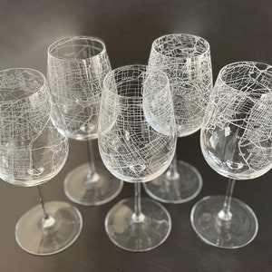 May include: Five clear glass wine glasses with a white city map design etched on the surface. The glasses are arranged in a semi-circle on a dark gray surface.