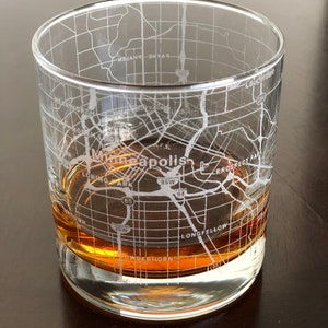 May include: A clear glass with a map of Minneapolis etched into the side. The glass is filled with a brown liquid.