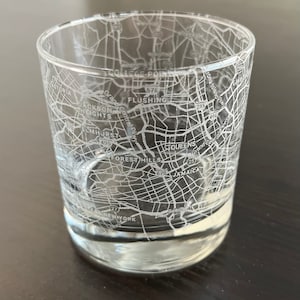 May include: A clear glass tumbler with an etched map of Queens, New York City. The map includes street names and highway numbers, such as Grand Central Parkway, Cross Island Parkway, and the 495 highway. The names of neighborhoods are also included, such as Flushing, Forest Hills, and Jamaica.