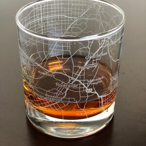 May include: A clear glass tumbler with a design etched into the side. The design is a map of Riverside, California, with street names and landmarks visible. The tumbler is filled with a brown liquid.