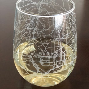 May include: A clear glass wine glass with a white etched design of the Washington, D.C. street map. The glass is filled with a light yellow liquid.