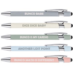May include: Five retractable ballpoint pens with chrome accents and black stylus tips. The barrels are matte gray, cream, sage green, light blue, and soft pink. Each pen features white text: BUNCO BABE, DICE DICE BABY, BUNCO IS MY CARDIO, ANOTHER LOST POINT, and BUNCO MATH IS DIFFERENT.