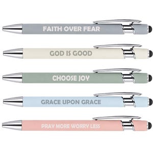 May include: A set of five retractable ballpoint pens with silver-toned metal clips and tips. The barrels are matte gray, cream, sage green, light blue, and soft pink, each featuring white text: FAITH OVER FEAR, GOD IS GOOD, CHOOSE JOY, GRACE UPON GRACE, PRAY MORE WORRY LESS.