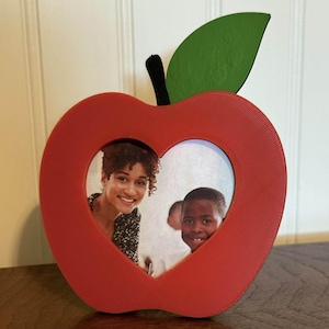 May include: Red apple-shaped picture frame with a heart-shaped opening. The frame has a green leaf and a black stem. The frame holds a photo of a smiling woman and a smiling child. The frame is on a wooden surface.
