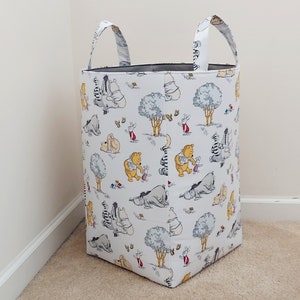 May include: White canvas laundry basket with two handles. The basket is decorated with a Winnie the Pooh print featuring Winnie the Pooh, Piglet, Tigger, Eeyore, and other characters from the Hundred Acre Wood.