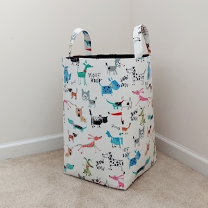 May include: A white fabric storage bin with black handles. The bin is decorated with a colorful pattern of dogs and the words "Woof Woof", "Bow Wow", and "Woof".