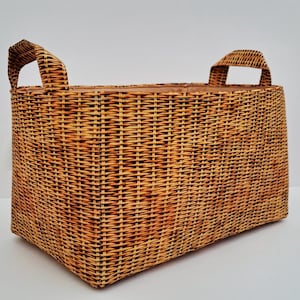 Puede incluir: Una cesta rectangular de mimbre tejido, de color marrón cálido, con dos asas arqueadas. Se aprecia la construcción de tejido apretado de la cesta. Las asas están integradas en el diseño, ofreciendo funcionalidad y atractivo visual. La cesta mide unos 40 cm de largo.