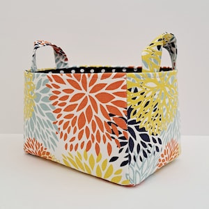 Könnte beinhalten: Ein rechteckiger Stoff-Aufbewahrungskorb mit zwei kleinen Griffen. Der Korb hat ein Blumenmuster in Orange, Gelb, Blau und Marine auf weißem Hintergrund. Das Innenfutter ist schwarz mit weißen Punkten.