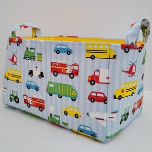Puede incluir: Un cesto de almacenamiento rectangular de tela con un fondo a rayas azules y blancas. El cesto está decorado con ilustraciones coloridas de varios vehículos, incluidos coches, camiones y autobuses. El interior es amarillo y tiene dos asas.