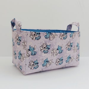 Peut inclure: Un panier de rangement en tissu lavande avec des poignées bleues et un motif répété de Stitch et Angel de Lilo & Stitch en train de se câliner.