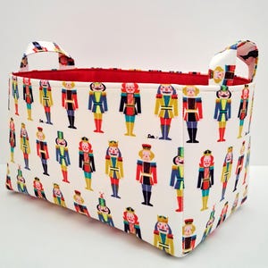 May include: A rectangular fabric storage basket with a white exterior patterned with colorful nutcracker figures. The interior is red, and the basket has two white handles with a patterned design. The basket is suitable for storing toys or other items.