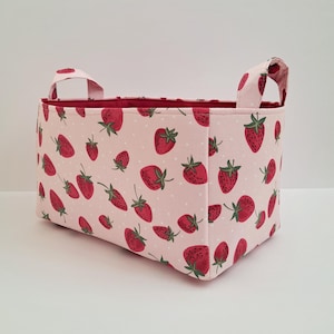 May include: A pink fabric storage basket with white polka dots and a strawberry print. The basket has two handles and is made of a soft, durable fabric.