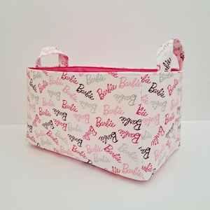 May include: A white fabric storage bin with pink and silver Barbie logos printed all over. The bin has two pink fabric handles.