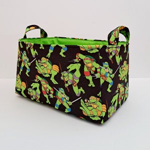 May include: A black and green fabric storage bin with a Teenage Mutant Ninja Turtles print. The bin has two handles and a green lining.