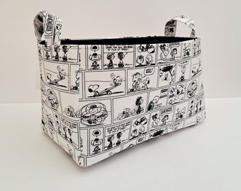 Puede incluir: Un cesto de almacenamiento rectangular con dos asas, con un patrón de cómic en blanco y negro. El interior es negro. El diseño del cómic incluye varios personajes y bocadillos.
