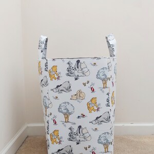 May include: A white laundry hamper with Winnie the Pooh and friends printed on it. The hamper has two handles and is standing on a beige carpet.