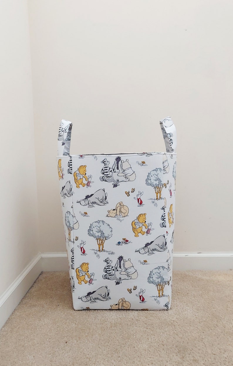 May include: A white laundry hamper with Winnie the Pooh and friends printed on it. The hamper has two handles and is standing on a beige carpet.