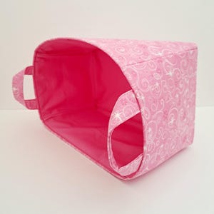 May include: Pink fabric storage bin with white swirl design and a pink lining. The bin has two handles.