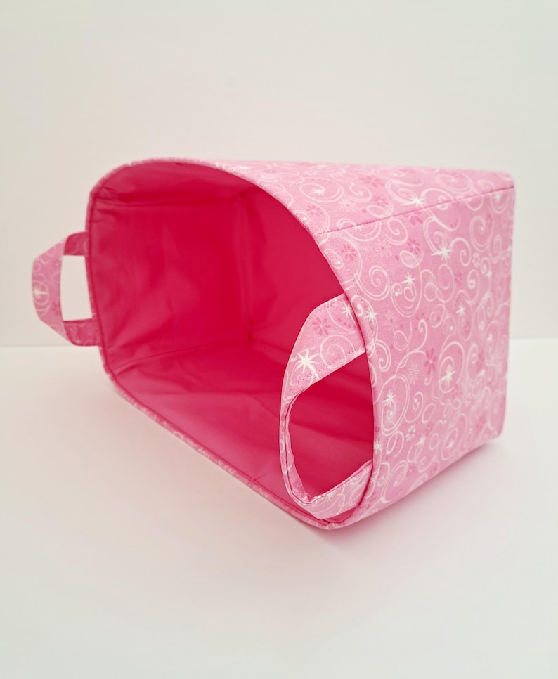 May include: Pink fabric storage bin with white swirl design and a pink lining. The bin has two handles.