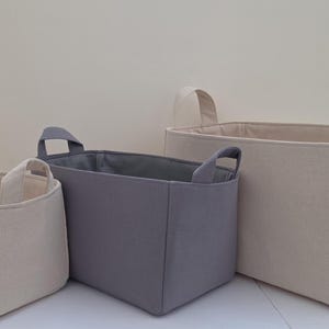 May include: Three rectangular fabric storage baskets with handles. The smallest and largest baskets are off-white, while the middle-sized basket is gray. Each basket features a structured design with reinforced side handles for easy carrying.